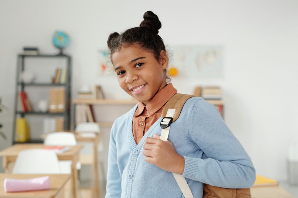 Smiling schoolgirl wearing a blue sweater stands in a classroom, ready for school.